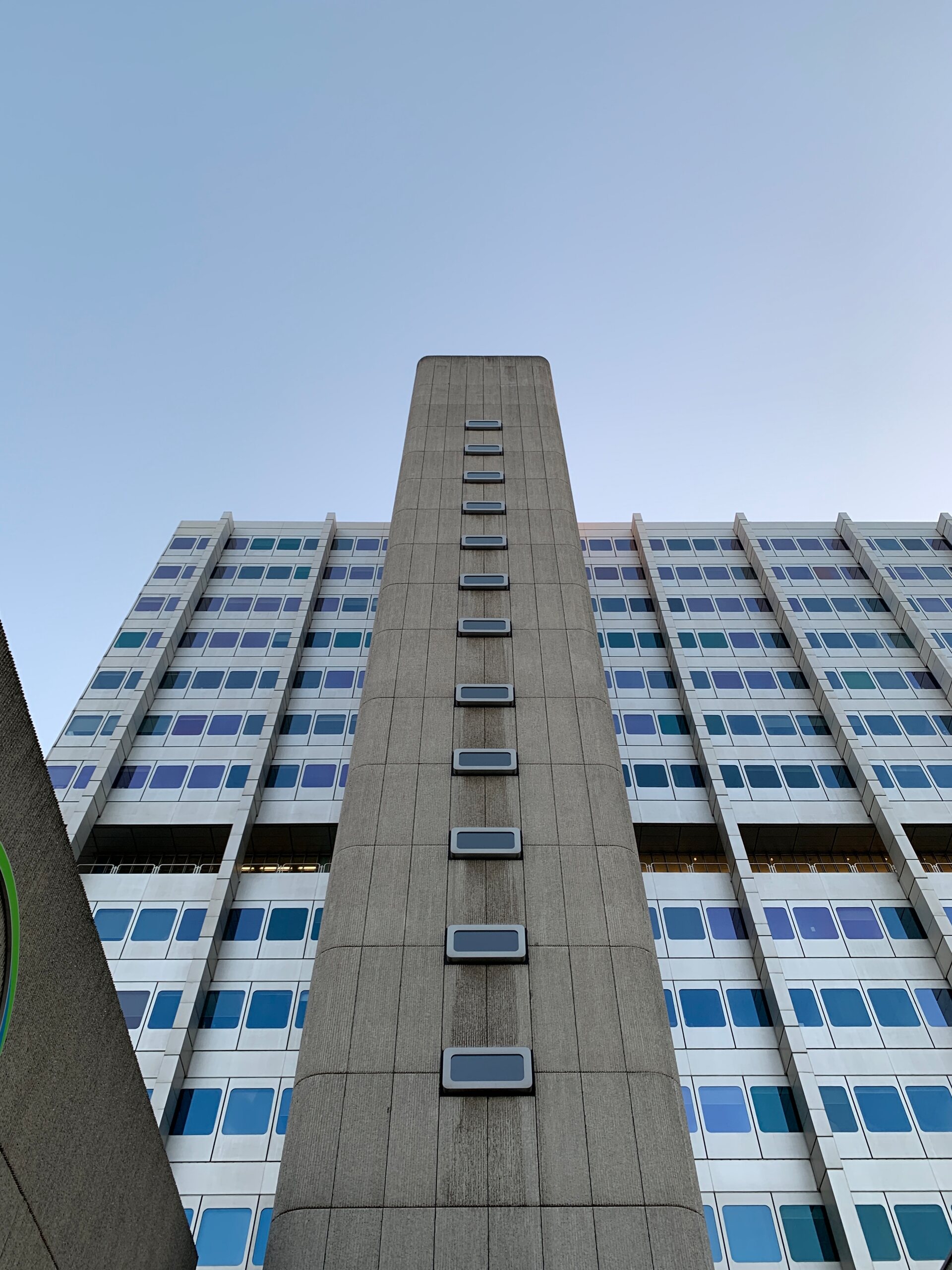 A concrete tower block seen from below, a blank shaft of small windows rising up its middle between wings of glass, against a clear pale sky.