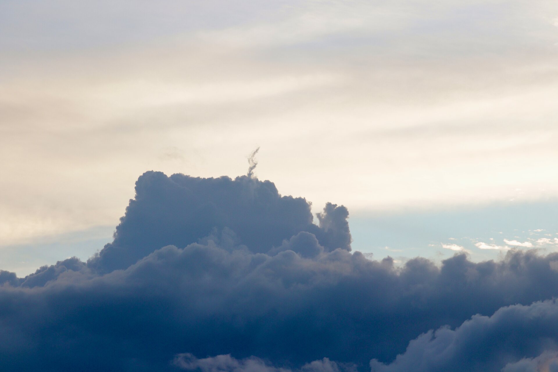 A tall dark cumulus cloud standing up out of a bank of cloud under a pale evening sky, a thin wisp curling up from its top.