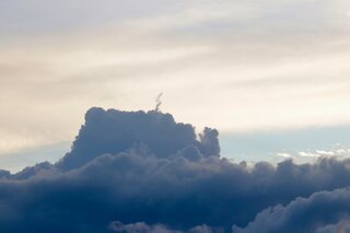 A tall dark cumulus cloud standing up out of a bank of cloud under a pale evening sky, a thin wisp curling up from its top.
